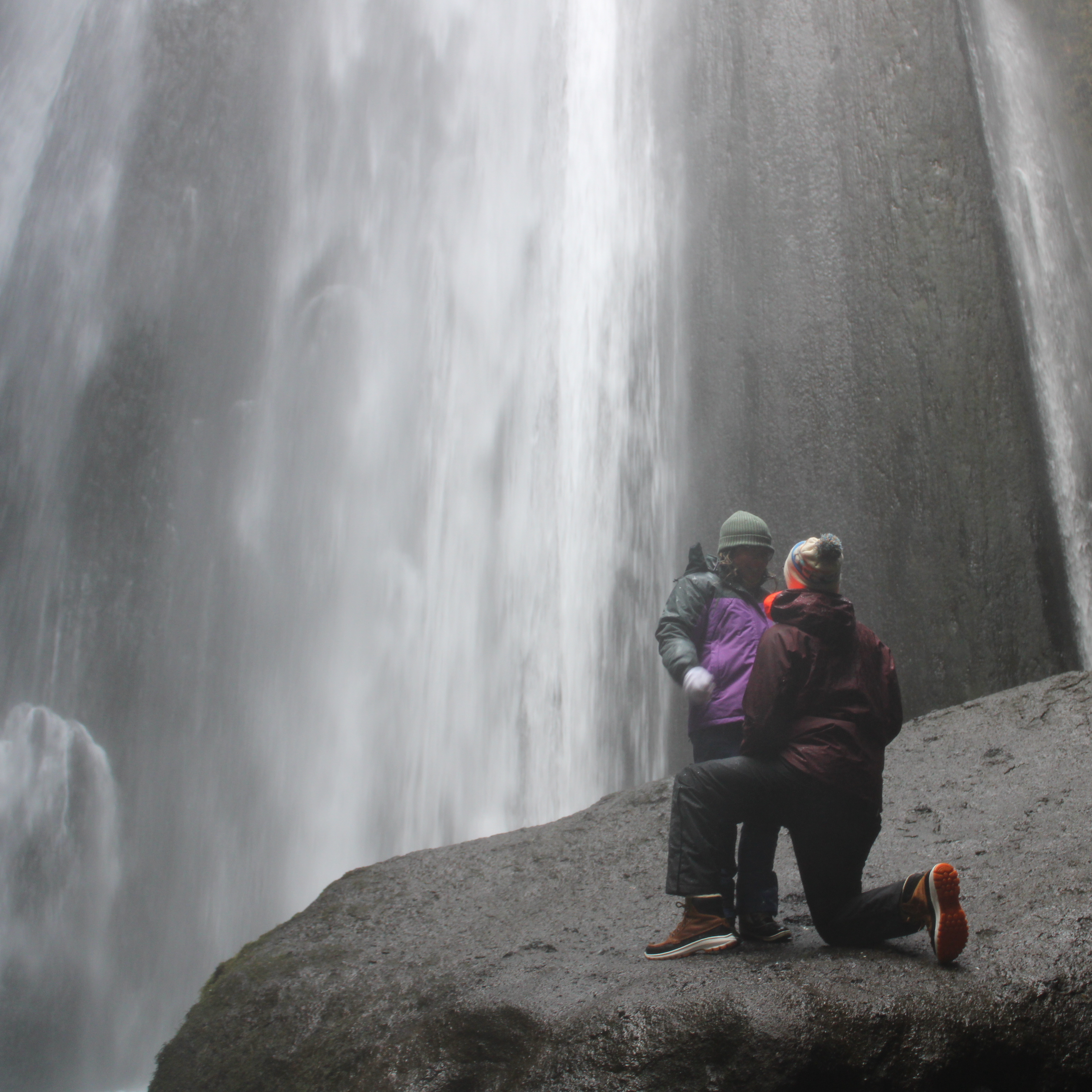 On top of a rock, in a cave, under a waterfall, is a very tall man proposing to a very tiny woman.