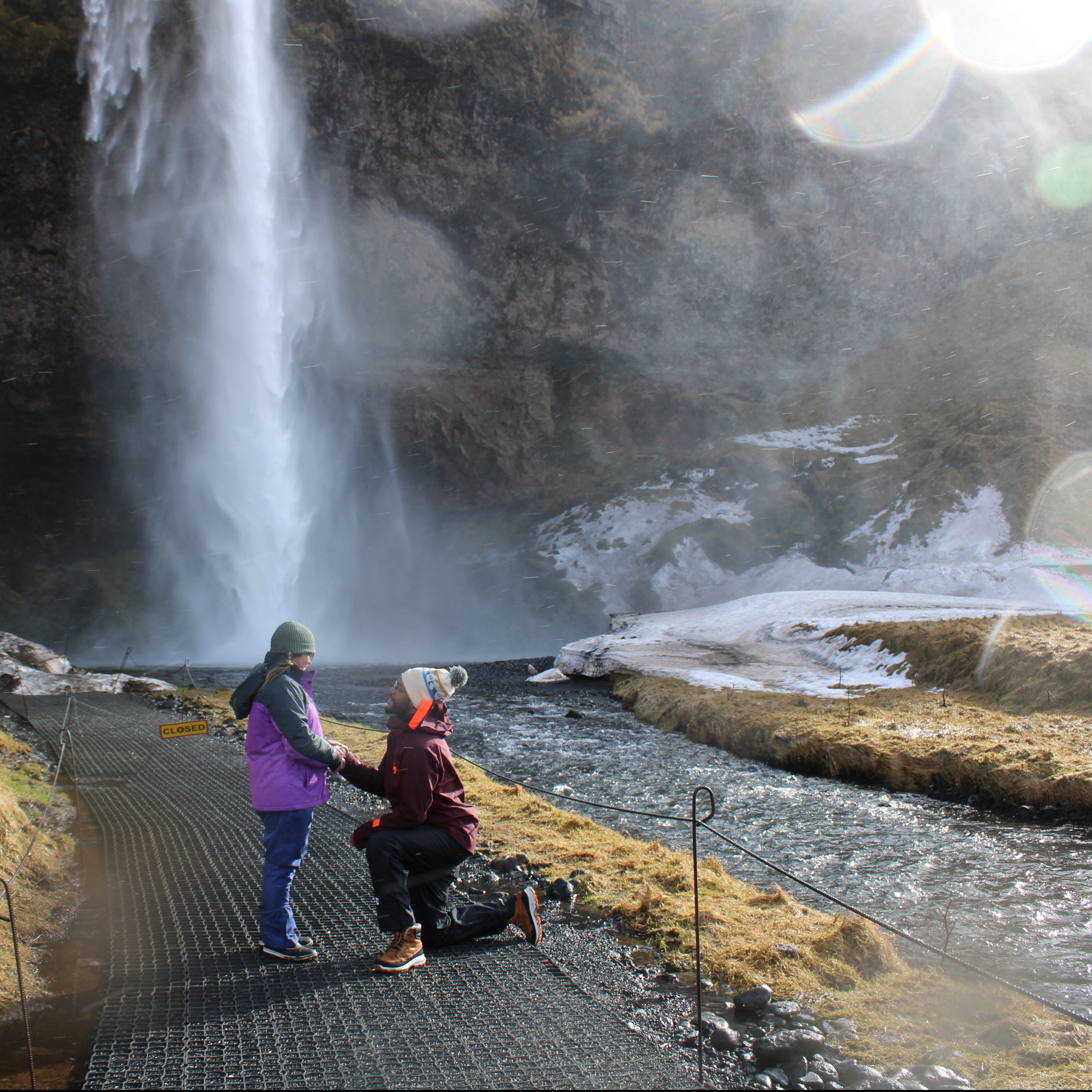 Beside the river that the waterfall feeds into is a man in red proposing to a girl in purple.