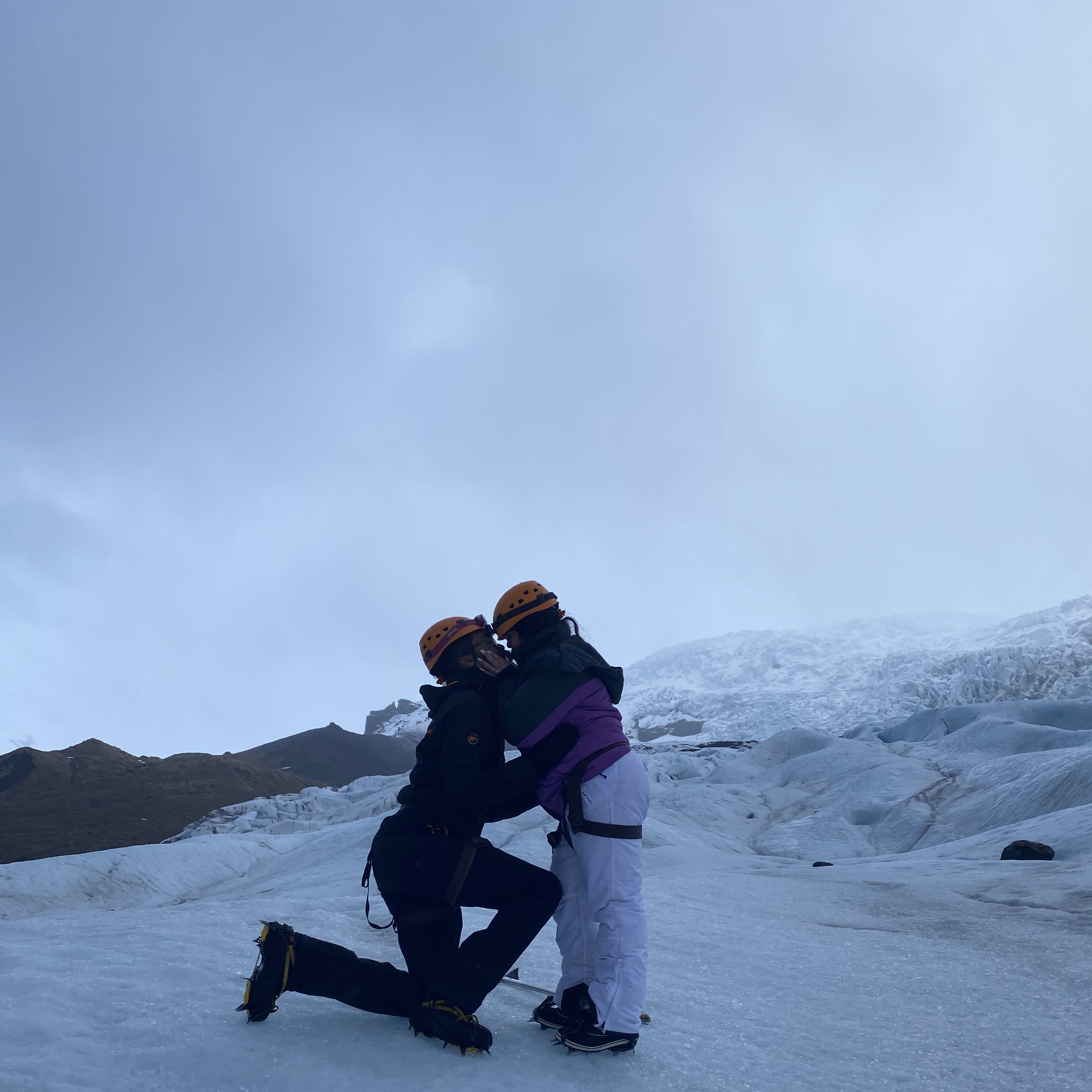 A man proposing to a girl who is kissing him while they both wear crampons standing on top of a Glacier.