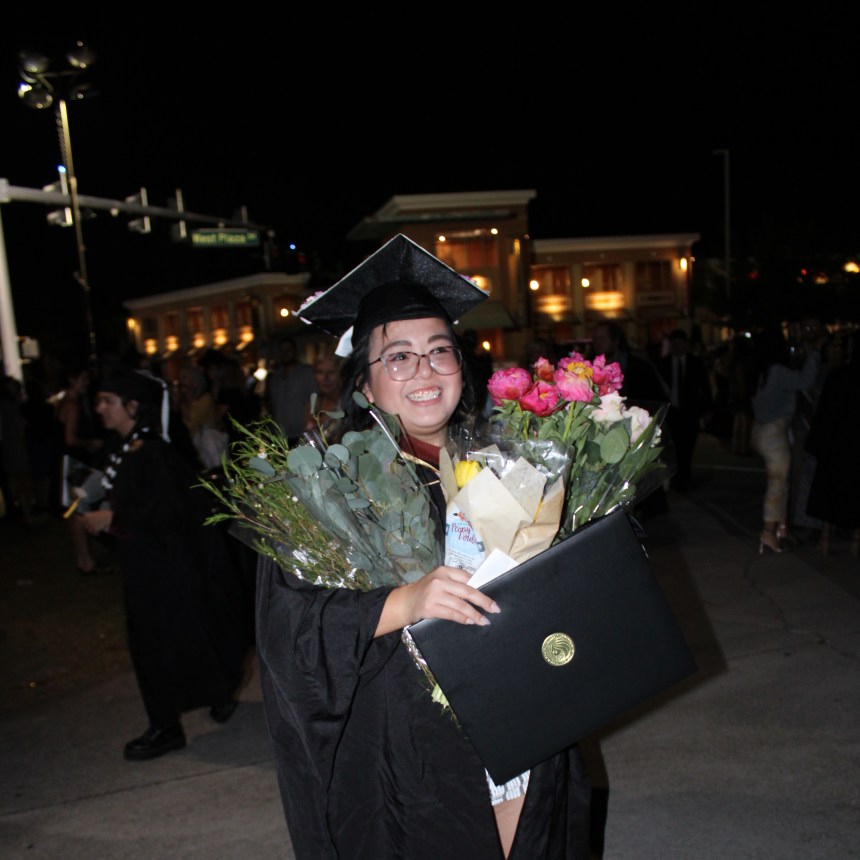 A short little girl staring away from the camera, big smile on her face, arms full of flowers and graduation memorabilia, probably sweating to death under her graduation robe.